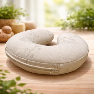 May include: A beige travel pillow with a U-shape design. The pillow features a decorative floral pattern and a zipper closure. The pillow is designed for neck support and comfort.