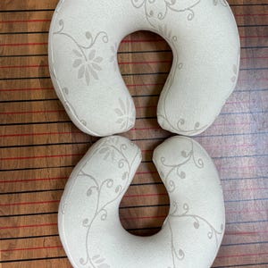 May include: Two U-shaped travel pillows in a cream-colored fabric featuring a subtle, light beige floral vine pattern. The pillows have a curved, ergonomic design intended for neck support, with a consistent textile texture across the surface.