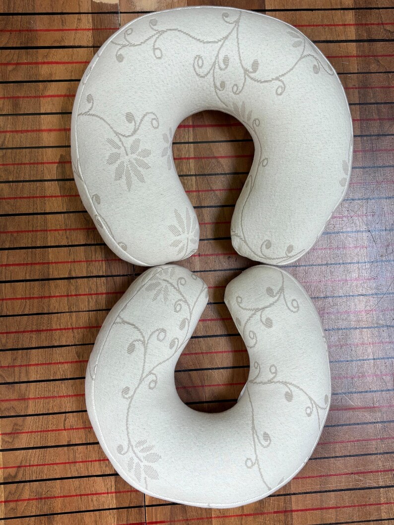 May include: Two U-shaped travel pillows in a cream-colored fabric featuring a subtle, light beige floral vine pattern. The pillows have a curved, ergonomic design intended for neck support, with a consistent textile texture across the surface.