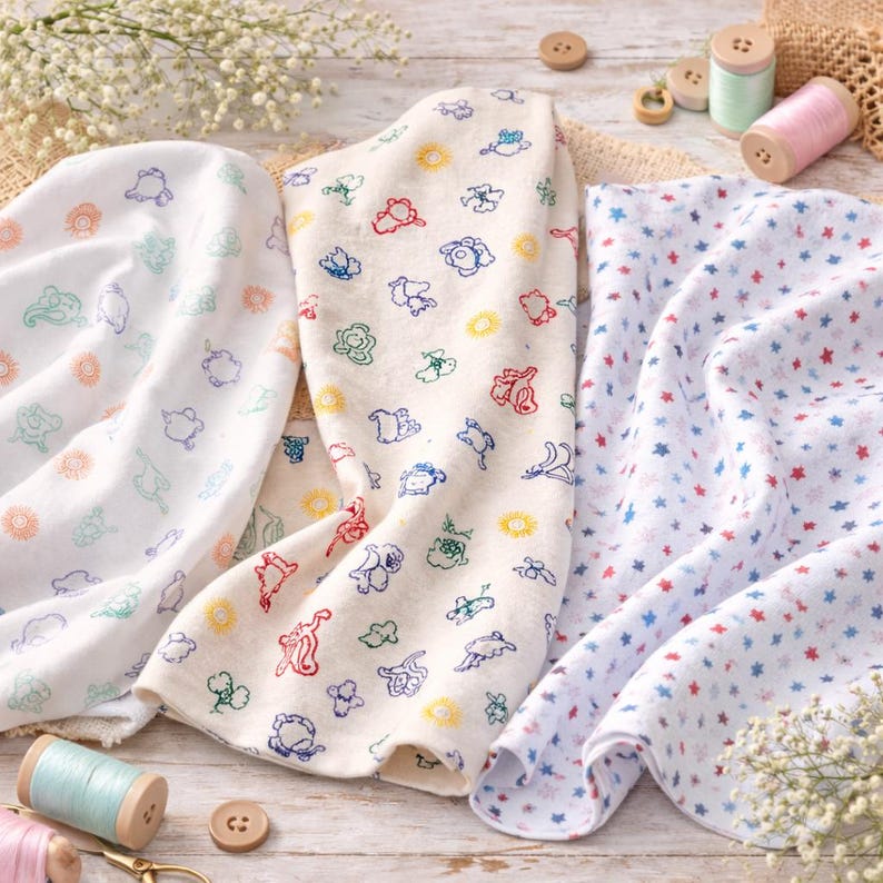 May include: Three folded baby blankets with embroidered designs. One is white with colorful animal shapes, another is white with blue, green, and orange cloud-like shapes, and the third is white with red and blue stars. Wooden spools of thread and buttons are in the background.
