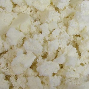 May include: Close-up of a pile of off-white foam pieces. The foam is irregularly shaped, with a porous texture and a slightly uneven surface. The pieces vary in size and are tightly packed together.