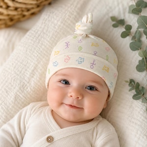 May include: A cream-colored baby hat with a knotted top, adorned with colorful, whimsical illustrations. The hat is worn by a smiling baby wearing a cream-colored onesie. The illustrations include fish, leaves, and other symbols.