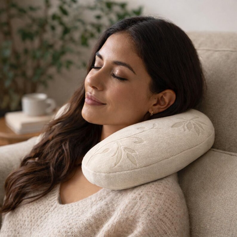 May include: A beige, crescent-shaped neck pillow featuring a subtle, tone-on-tone embroidered leaf pattern. The pillow has a soft, textured fabric surface and is designed to provide support around the neck area while resting against a chair.