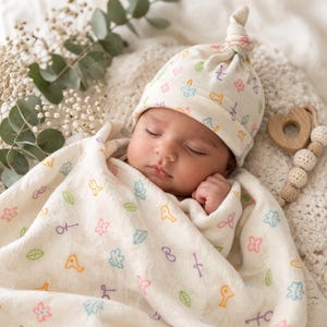 May include: A sleeping baby wrapped in a cream-colored blanket and wearing a matching hat, both adorned with colorful, whimsical illustrations. A wooden teething toy rests nearby. The scene evokes a sense of comfort and tranquility.