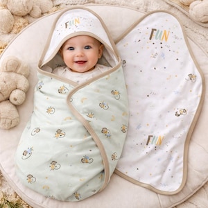 May include: A baby wrapped in a light green and white swaddle blanket with a matching hooded towel. The blanket features a pattern of monkeys, moons, and stars. The word "FUN" is printed on the hood and blanket in colorful letters. The edges are trimmed in a tan color.