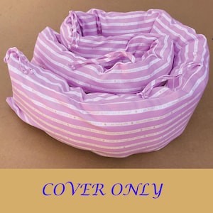 May include: A pink and white striped cot bumper cover. The cover is made of fabric and has a drawstring closure. The text "COVER ONLY" is printed on the bottom of the image.