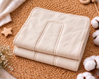 May include: A rectangular, off-white cloth diaper insert featuring a multi-layered design with reinforced stitching along the edges and a central panel. The fabric has a soft, matte texture and is folded neatly into a compact, layered stack.