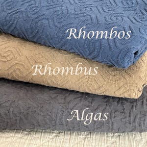 May include: Three folded pieces of textured fabric stacked vertically. The top piece is blue with the text Rhombos, the middle piece is tan with the text Rhombus, and the bottom piece is dark grey with the text Algas printed in white.
