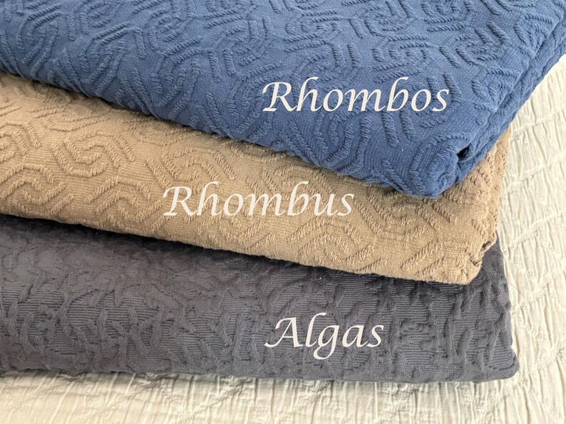 May include: Three folded pieces of textured fabric stacked vertically. The top piece is blue with the text Rhombos, the middle piece is tan with the text Rhombus, and the bottom piece is dark grey with the text Algas printed in white.