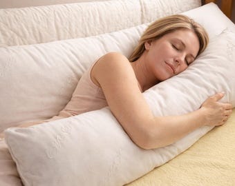 May include: A long, rectangular off-white body pillow with a soft, textured fabric cover. The pillow features an embossed logo pattern across its surface. It is designed to support the full length of the body while resting in a horizontal position.