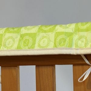 May include: A lime green quilted crib rail cover with a repeating lion face pattern. The cover is trimmed with a cream-colored fabric and tied to the crib with white ribbons. The crib is made of wood.