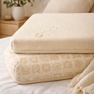 May include: A rectangular beige pillow with a smooth surface featuring several small water droplets on top. Beneath it sits a second rectangular pillow in a lighter cream shade, decorated with a repeating pattern of tan geometric shapes.