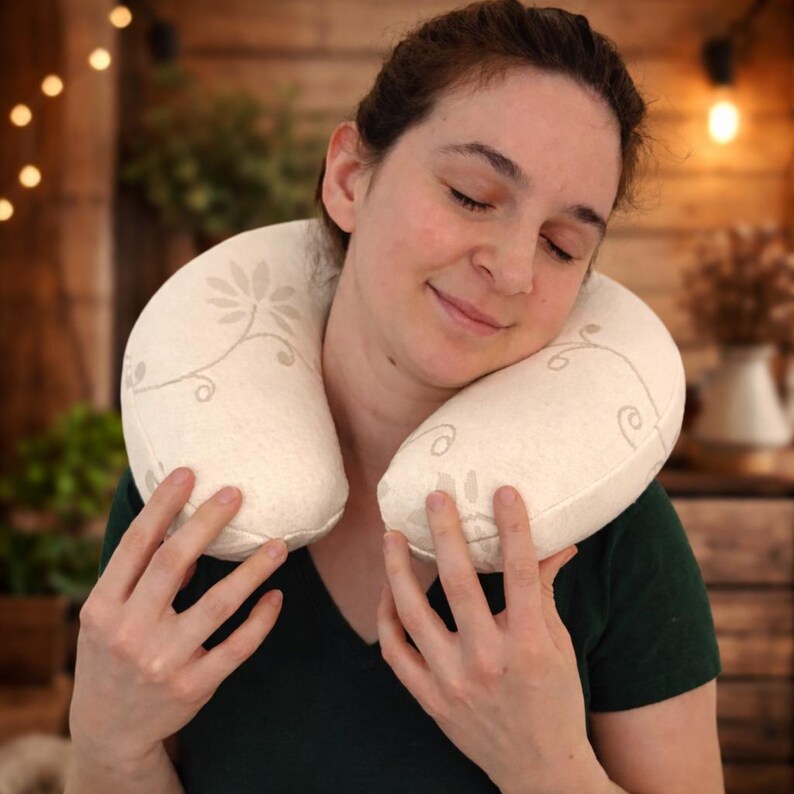 May include: A U-shaped travel neck pillow in an off-white or cream color, featuring a delicate, light brown floral vine pattern printed across the fabric surface. The pillow is designed to wrap around the neck for support and comfort.
