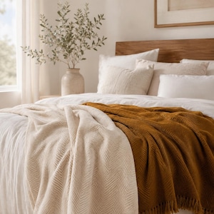 May include: A bed featuring a white duvet, a cream-colored herringbone throw blanket, and a mustard-yellow herringbone throw blanket with fringe edges. The bed is layered with multiple white and beige pillows against a wooden headboard.