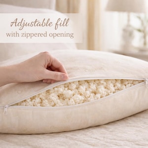 May include: A beige pillow with a zippered opening, revealing a fluffy, white interior. The text "Adjustable fill with zippered opening" is displayed above the pillow. The pillow is on a bed.