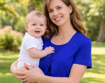 Peut inclure: Une femme en haut bleu tient un bébé en barboteuse blanche. Les deux sourient. L'arrière-plan est une scène extérieure verte floue avec des arbres.