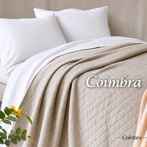 May include: A beige quilted bedspread with a diamond pattern and the text Coimbra printed on it. The bedspread is layered over white bed sheets and two white pillows. A small portion of a peach-coloured knit blanket is visible in the corner.