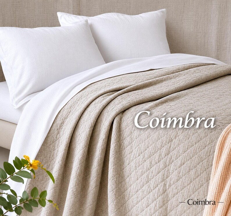 May include: A beige quilted bedspread with a diamond pattern and the text Coimbra printed on it. The bedspread is layered over white bed sheets and two white pillows. A small portion of a peach-coloured knit blanket is visible in the corner.