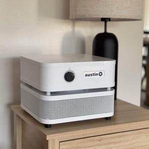 May include: White air purifier with a black grill and a black knob on the top. The air purifier has a sticker that says "austin it" on the side. The air purifier is sitting on a wooden table.