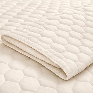 May include: A cream-colored quilted blanket featuring a repeating hexagonal pattern across the entire surface. The fabric has a soft, textured appearance with visible stitching defining each hexagon shape, and one corner is folded over.