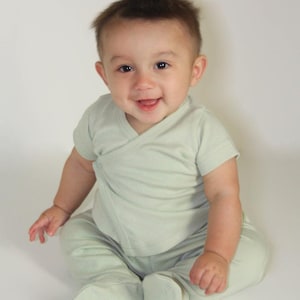 May include: A smiling baby wearing a light green wrap-style top and matching footed pants. The baby is sitting and looking directly at the camera. The outfit appears to be made of soft, comfortable material.