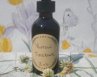 May include: A dark amber glass bottle of Yarrow Tincture with a black dropper top. The label reads "Yarrow Tincture 2 oz" in handwritten script. The bottle is surrounded by small white flowers and rests on a floral patterned fabric.
