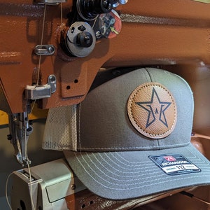 May include: A brown leather baseball cap with a brown leather star patch on the front. The cap is sitting on a brown leather sewing machine. The machine has a gold plate that reads "Leather Machine Company".
