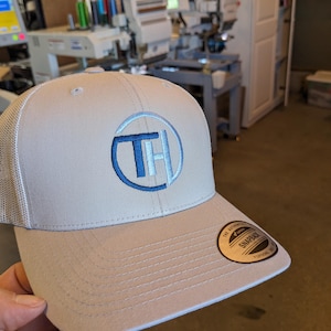 May include: A light gray trucker hat with a blue and white embroidered logo of the letters "TH" inside a circle. The hat has a black "The Authentic Snapback" label on the back.