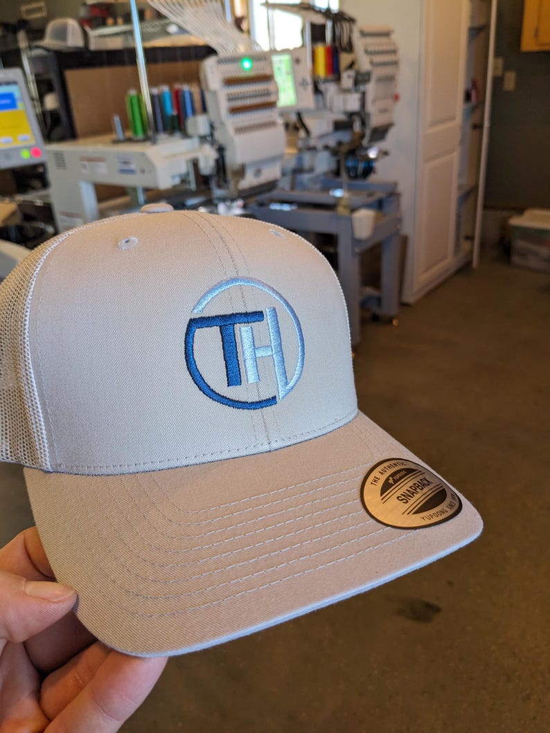 May include: A light gray trucker hat with a blue and white embroidered logo of the letters "TH" inside a circle. The hat has a black "The Authentic Snapback" label on the back.