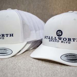 May include: Two white mesh trucker hats with a navy blue embroidered logo that says "Stallworth Grand Blvd" and a navy blue "S" in a circle. The hats have a black "The Authentic Snapback" label on the underside of the brim.