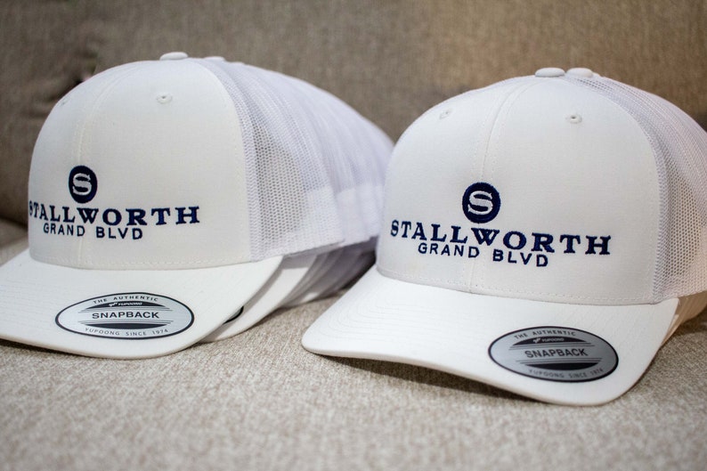 May include: Two white mesh trucker hats with a navy blue embroidered logo that says "Stallworth Grand Blvd" and a navy blue "S" in a circle. The hats have a black "The Authentic Snapback" label on the underside of the brim.
