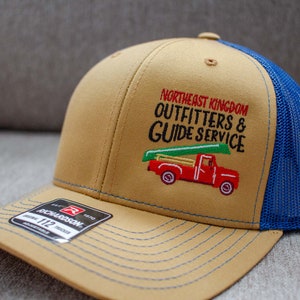 May include: A tan and blue trucker hat with an embroidered logo that says "Northeast Kingdom Outfitters & Guide Service" and a red pickup truck with a canoe on top. The hat is made by Richardson.
