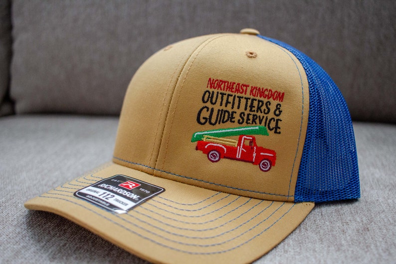 May include: A tan and blue trucker hat with an embroidered logo that says "Northeast Kingdom Outfitters & Guide Service" and a red pickup truck with a canoe on top. The hat is made by Richardson.