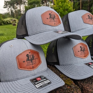 May include: Grey trucker hats with black mesh backs and brown leatherette patches. The patches feature a compass design with a feather and the letters N, W, and S. The hats are adjustable and have a Richardson tag.