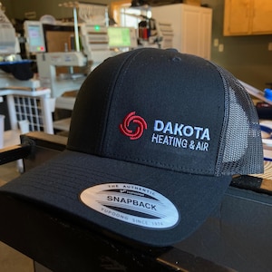 May include: Black and gray trucker hat with a red and white logo that says "Dakota Heating & Air".