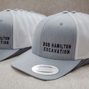 May include: Two gray and white trucker hats with black embroidered text that reads "Bob Hamilton Excavation". The hats have a mesh back and a snap closure.