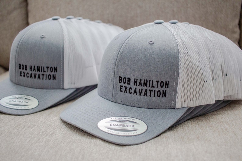 May include: Two gray and white trucker hats with black embroidered text that reads "Bob Hamilton Excavation". The hats have a mesh back and a snap closure.