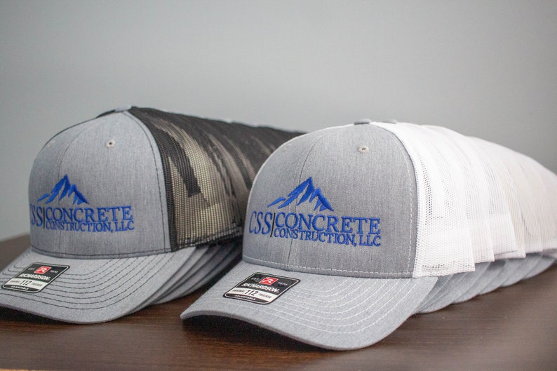 May include: A stack of gray and white trucker hats with a black mesh back. The hats have a blue embroidered logo that says "CSS Concrete Construction, LLC" with a mountain graphic.
