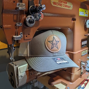 Puede incluir: Una gorra de b&eacute;isbol gris con parte trasera de malla beige y un parche circular de cuero marr&oacute;n claro con un logotipo de estrella negra. La gorra tiene una pegatina Richardson 112 en la visera y est&aacute; sobre una m&aacute;quina de coser industrial marr&oacute;n.