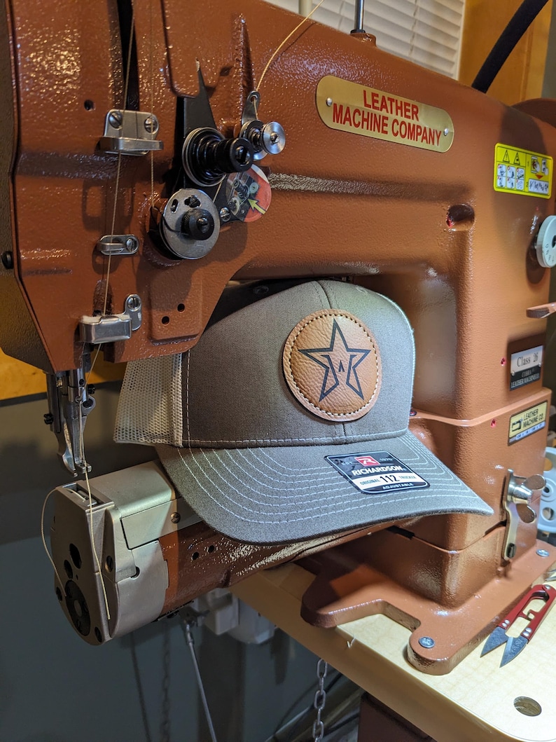 Puede incluir: Una gorra de b&eacute;isbol gris con parte trasera de malla beige y un parche circular de cuero marr&oacute;n claro con un logotipo de estrella negra. La gorra tiene una pegatina Richardson 112 en la visera y est&aacute; sobre una m&aacute;quina de coser industrial marr&oacute;n.