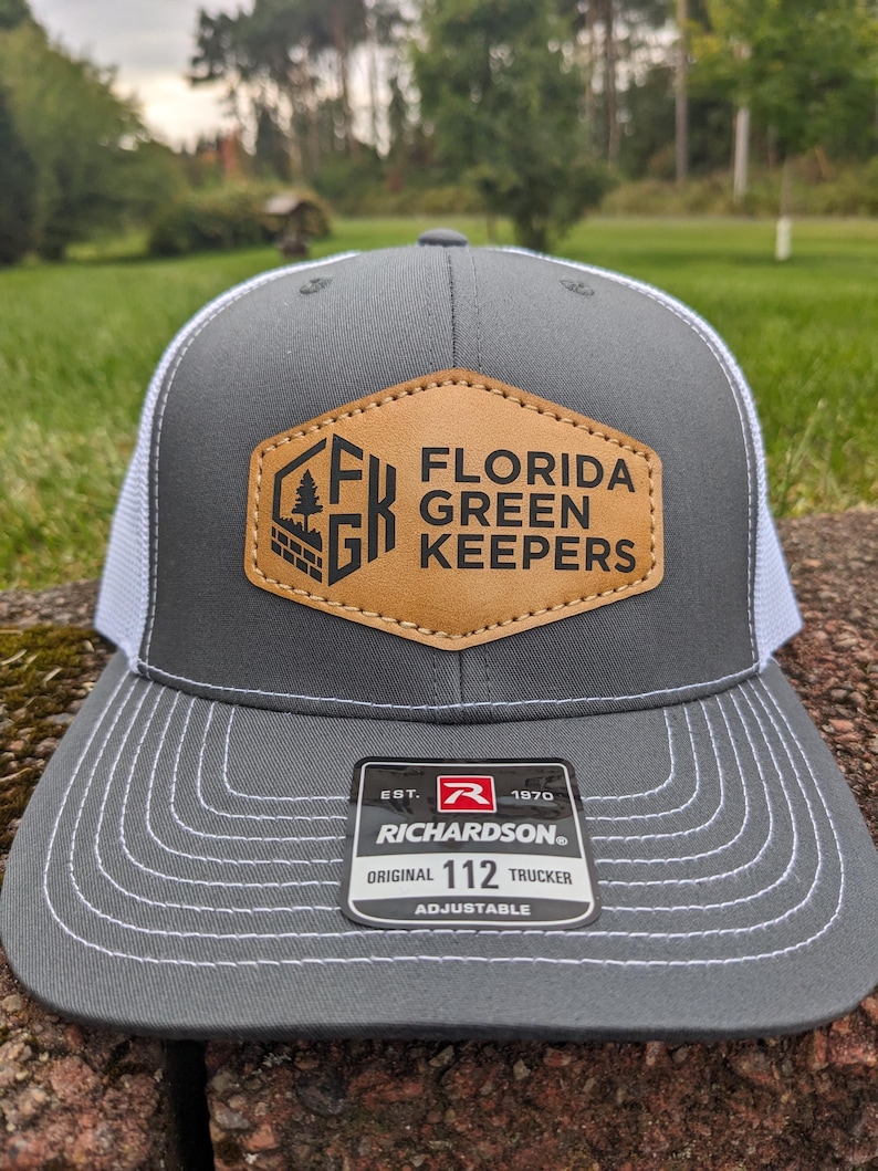 Puede incluir: Una gorra trucker gris y blanca con un parche hexagonal de cuero sint&eacute;tico color canela en la parte frontal. El parche muestra el texto FLORIDA GREEN KEEPERS y un logotipo con un &aacute;rbol. Una etiqueta negra en la visera dice EST. 1970 RICHARDSON 112 TRUCKER ADJUSTABLE.
