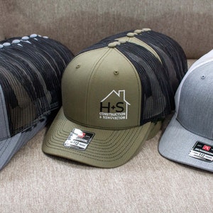 May include: Three trucker hats with black mesh back panels. The hats are gray, green, and white. Each hat has a logo embroidered on the front that says "H+S Construction & Renovation".