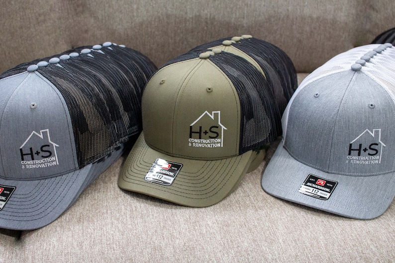 May include: Three trucker hats with black mesh back panels. The hats are gray, green, and white. Each hat has a logo embroidered on the front that says "H+S Construction & Renovation".