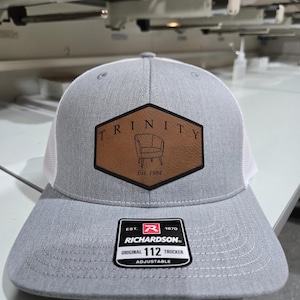 Puede incluir: Una gorra trucker gris claro con parte trasera de malla blanca y un parche de cuero hexagonal marr&oacute;n en el frente. El parche presenta el texto TRINITY, una ilustraci&oacute;n de una silla y Est. 1984. Una pegatina negra en la visera dice RICHARDSON 112.