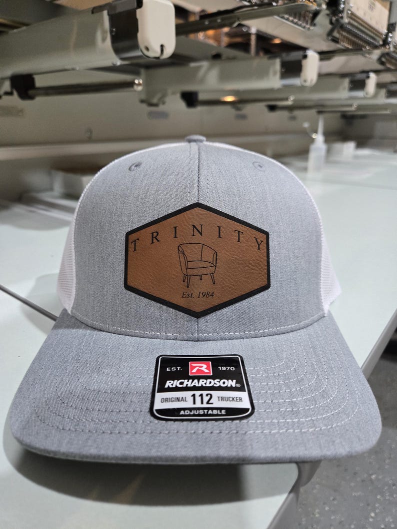 Puede incluir: Una gorra trucker gris claro con parte trasera de malla blanca y un parche de cuero hexagonal marr&oacute;n en el frente. El parche presenta el texto TRINITY, una ilustraci&oacute;n de una silla y Est. 1984. Una pegatina negra en la visera dice RICHARDSON 112.