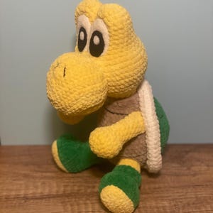 May include: A handmade crochet Koopa Troopa plush toy. The plush features a yellow head and limbs, a green shell, and green feet. The Koopa Troopa has black eyes and a white shell trim. The toy is sitting on a wooden surface.