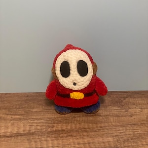 May include: A handmade plush toy of a Shy Guy character, featuring a red hooded outfit, a white mask with black eye holes, and blue shoes. The toy has a yellow belt and brown ear details. The plush is made of soft, textured yarn.