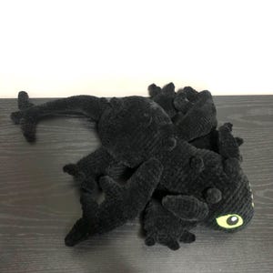 May include: Black crocheted plush toy dragon with green eyes. The dragon is lying on its side with its wings spread out.