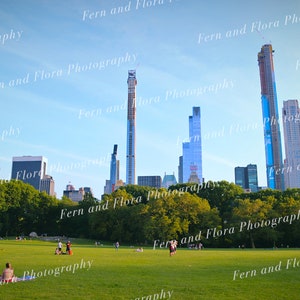 May include: A view of the New York City skyline from Central Park, with several tall buildings, including a skyscraper under construction, visible in the distance.  People are relaxing on the green grass in the foreground.