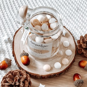 May include: A glass jar filled with small white and beige scented wax melts, labeled "Aromelyn Pixie Dust Handmade Soy Sconies Poured in Britain www.aromelynstudio.com". The jar is on a wooden slice with a wooden spoon and pine cones around it.